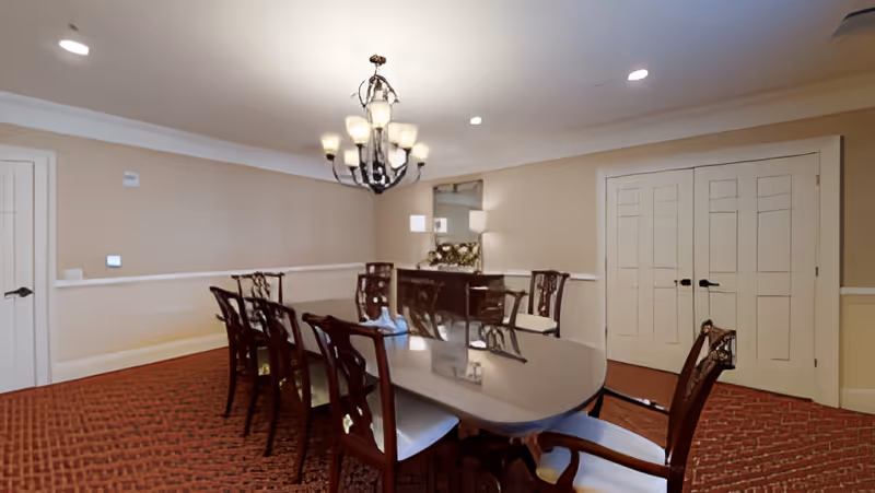 A formal dining room with a long polished wooden table surrounded by eight wooden chairs with cushioned seats. A chandelier with multiple lights hangs above the table. The room has beige walls, a red patterned carpet, a sideboard with decorative items, and a large mirror above it. There are two closed white double doors on one side and a single white door on another.