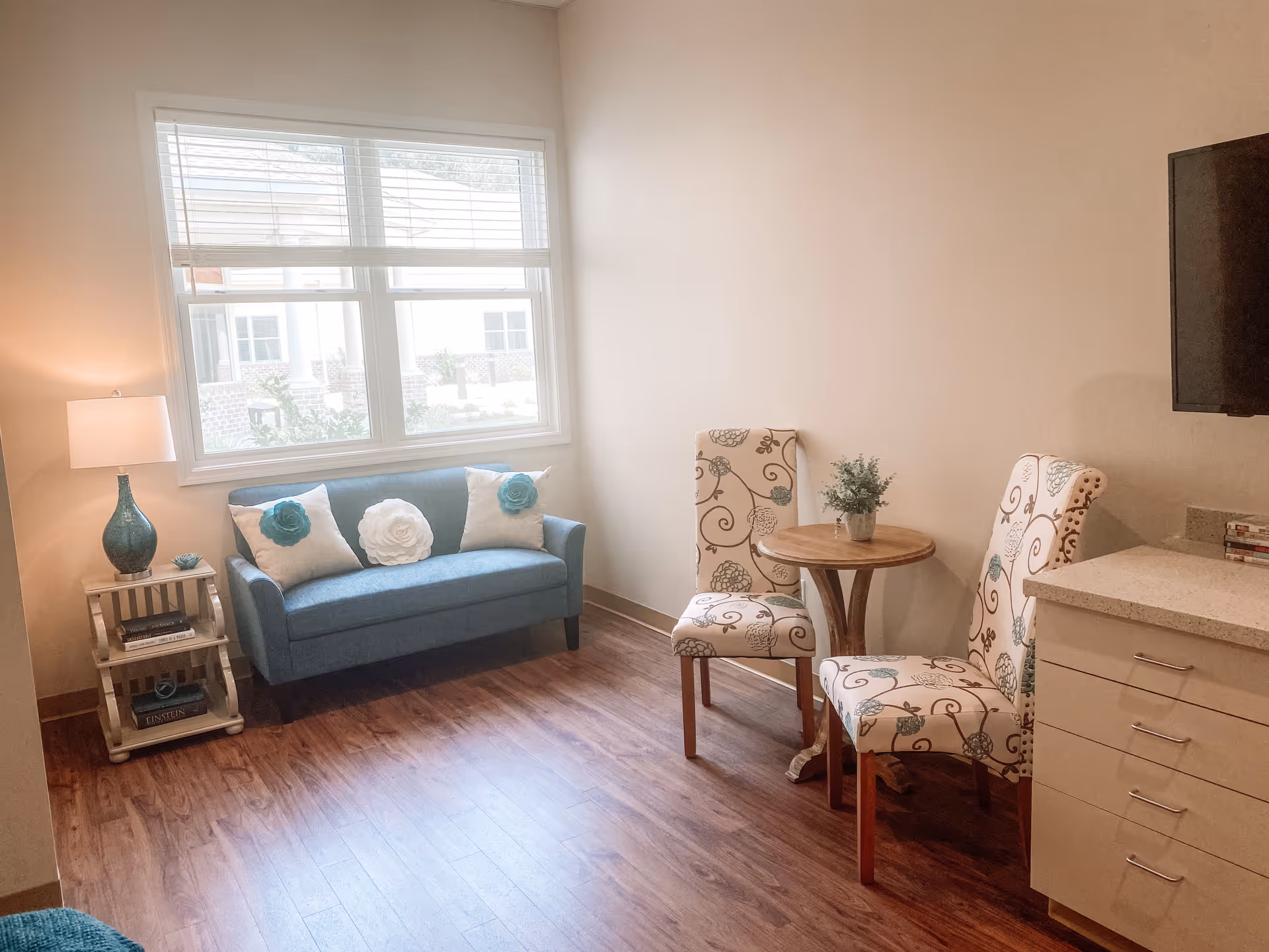 Small living room with a teal loveseat under a window, two patterned chairs around a round table, a side lamp and a wall-mounted TV.