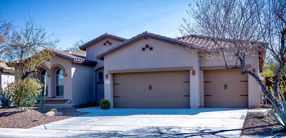 Exterior view of a single-story residential building with a three-car garage, beige stucco walls, and a tiled roof. The front yard features desert landscaping with rocks, small bushes, cacti, and leafless trees under a clear blue sky.