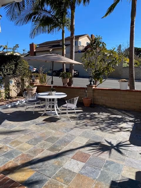 Outdoor patio area with a round white table and three white chairs under a large beige umbrella. Several potted plants and palm trees surround the patio, with a residential street and houses visible in the background under a clear blue sky.