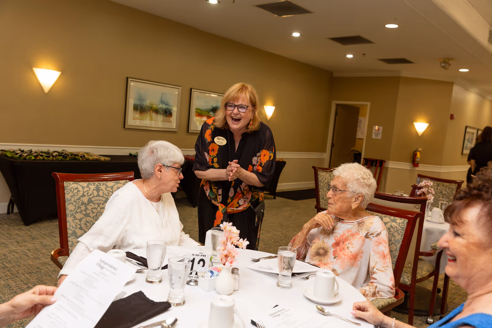 A group of elderly women sitting around a dining table in a senior living facility, engaging in conversation with a smiling staff member standing beside them. The table is set with menus, glasses, and cups, and the room has warm lighting with framed artwork on the walls.