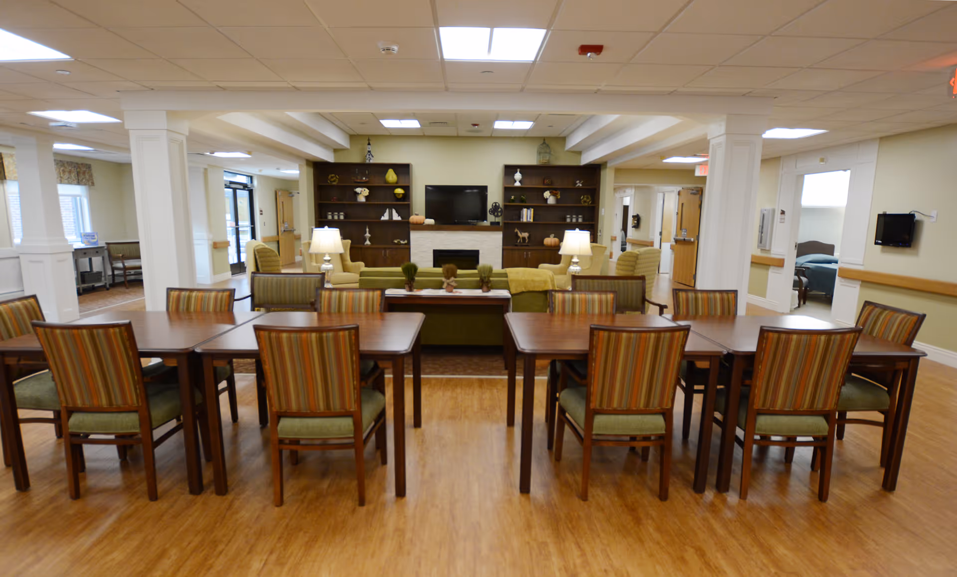 A spacious common area in a senior living facility featuring multiple wooden tables with striped cushioned chairs arranged neatly. In the background, there is a cozy seating area with green sofas, armchairs, two table lamps, and a fireplace with built-in shelves on either side holding decorative items. The room has a light-colored ceiling with recessed lighting and a wood floor.