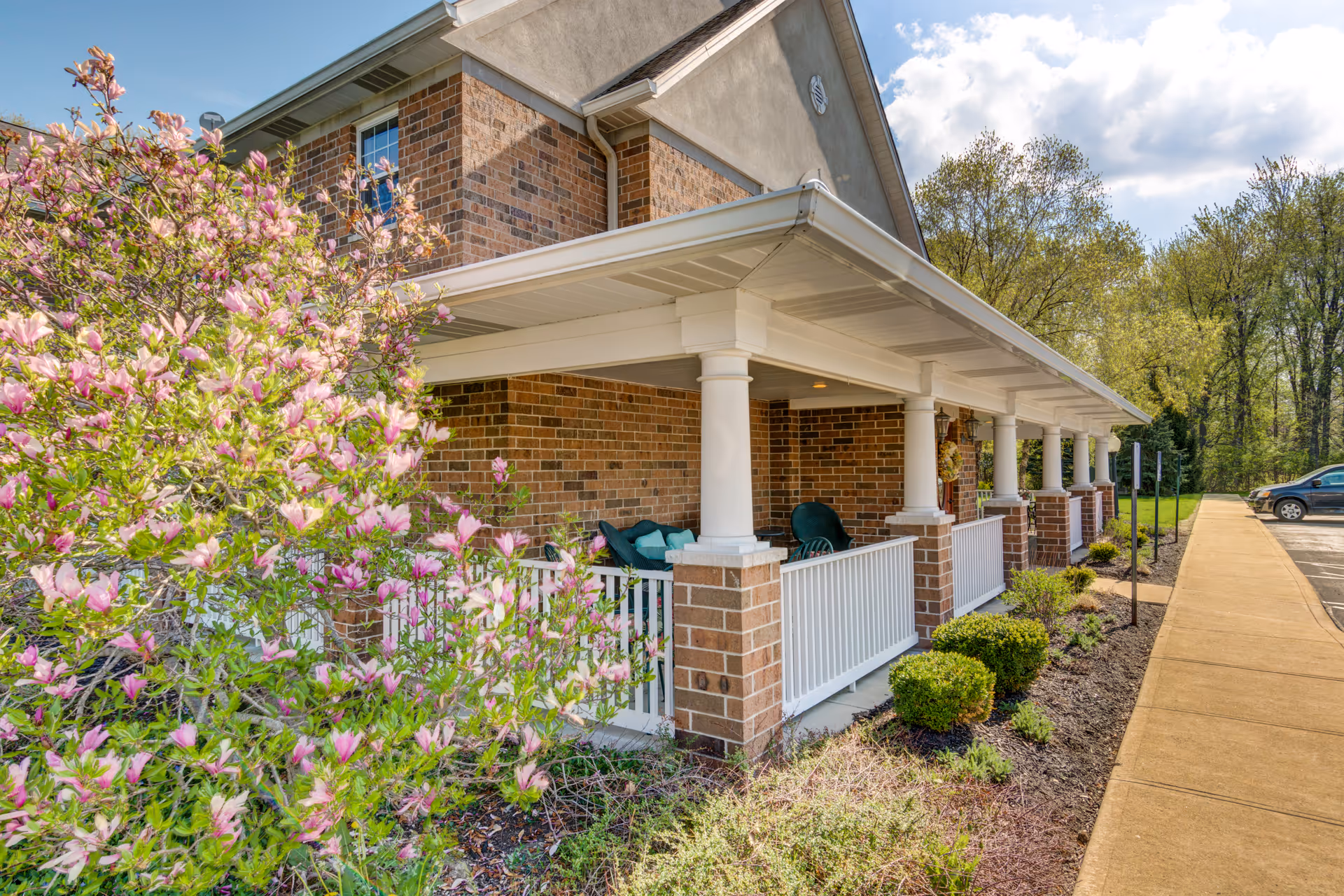 Exterior view of a brick building with a covered porch supported by white columns. The porch has green chairs and is surrounded by landscaped bushes and a blooming pink flower tree. A sidewalk and parking area are visible to the right, with trees in the background under a partly cloudy sky.