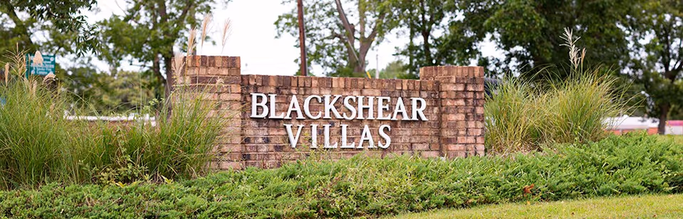 A brick sign with white letters reading 'Blackshear Villas' surrounded by green bushes and tall grass, with trees and a road in the background.