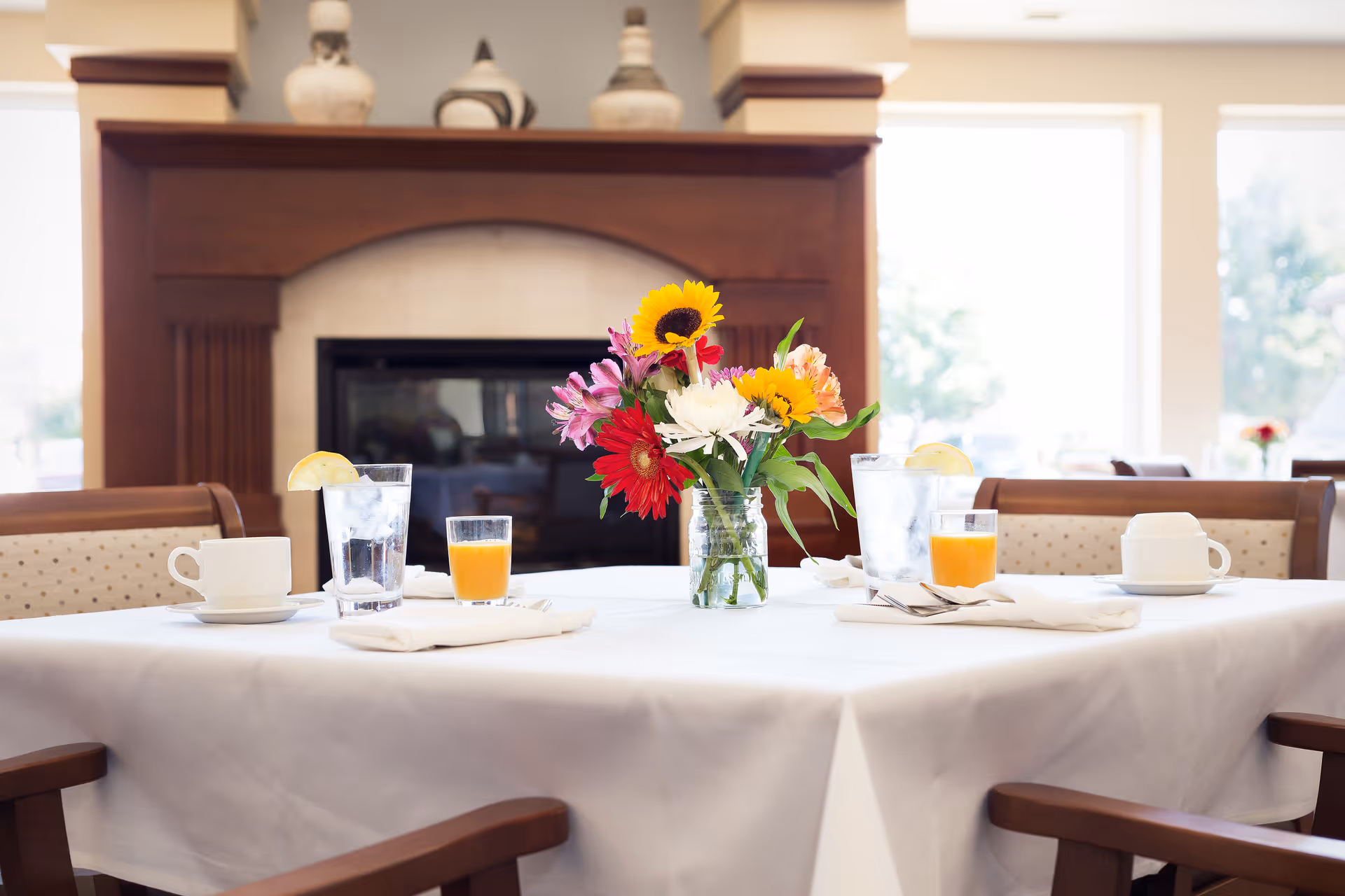 A dining table set with a white tablecloth, two glasses of water with lemon slices, two glasses of orange juice, two white coffee cups with saucers, and a vase of colorful flowers in the center. The background features a wooden fireplace and large windows letting in natural light.