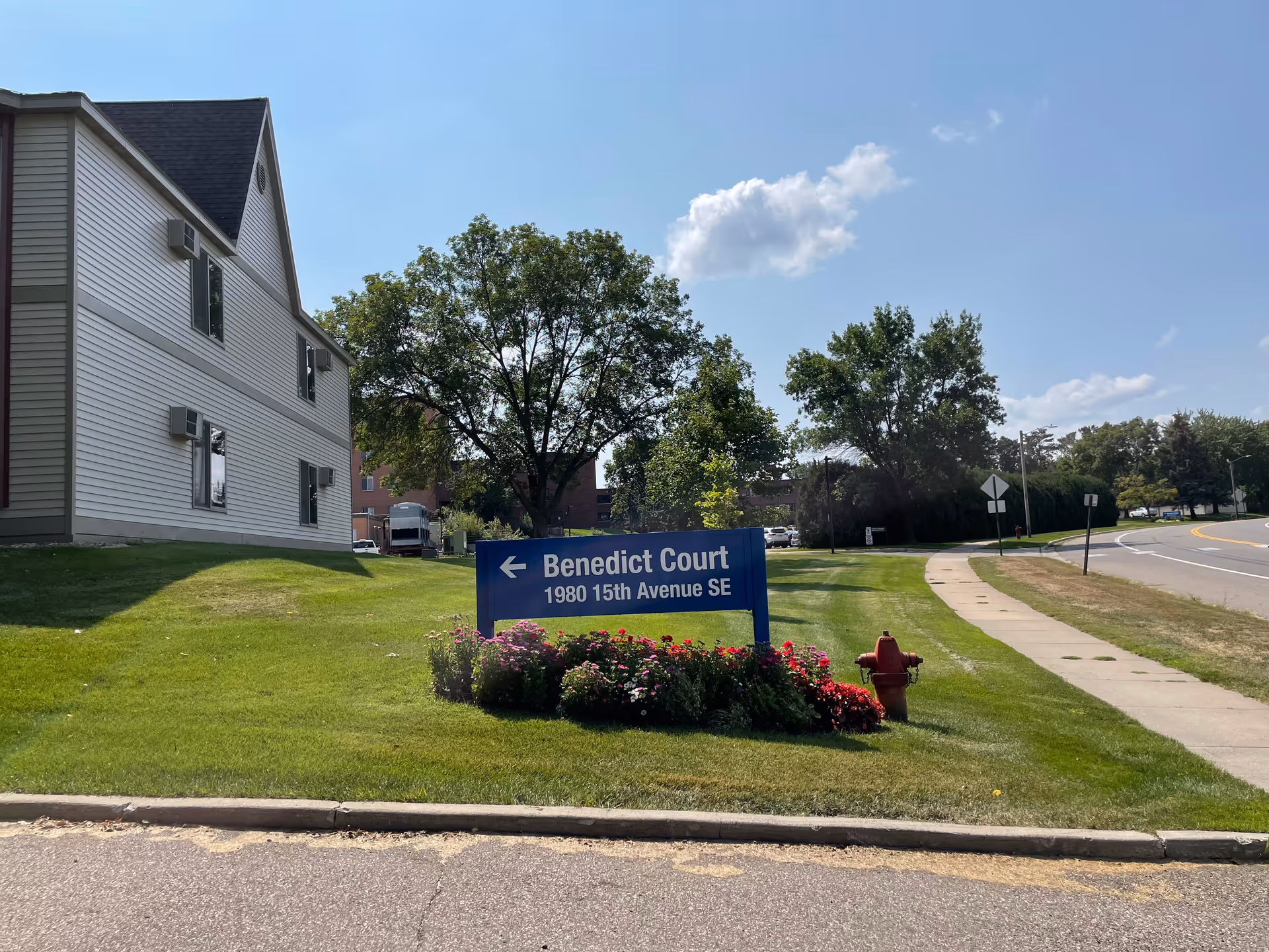 Blue sign reading "Benedict Court 1980 15th Avenue SE" on a grassy lawn in front of a light-colored building and a tree-lined street.