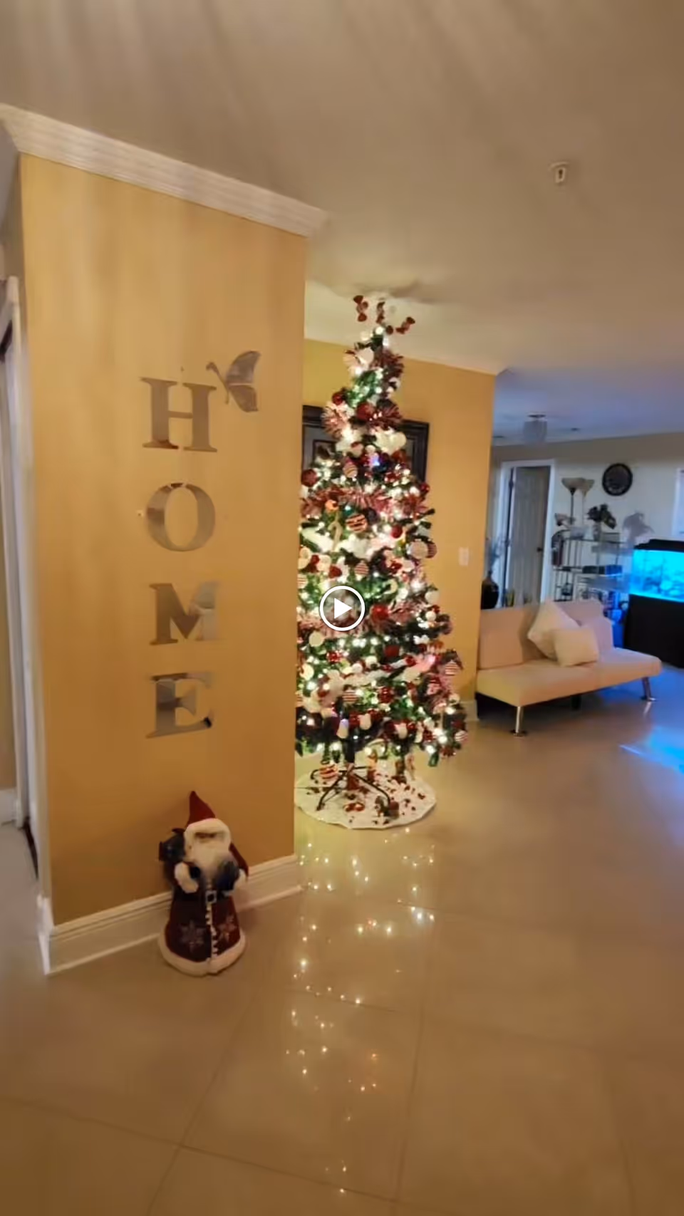 Interior view of a living room area decorated for Christmas with a lit Christmas tree, a small Santa Claus figure on the floor, and a wall decoration spelling the word HOME with a butterfly accent. There is a beige couch with pillows and a fish tank in the background.