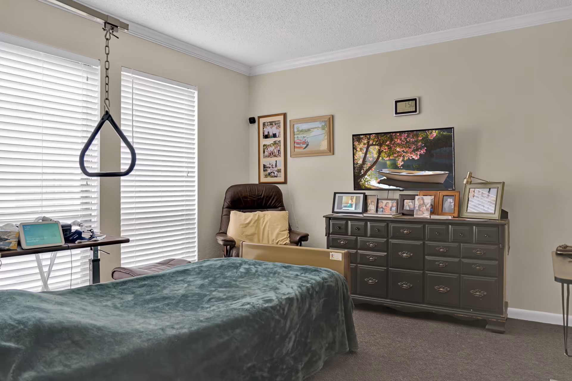 A cozy bedroom in a memory care facility featuring a bed with a green blanket, a brown recliner chair with a yellow pillow, a black dresser with multiple framed photos and a TV displaying a boat on a lake with blooming trees. The room has two large windows with white blinds and a trapeze bar hanging above the bed for assistance.