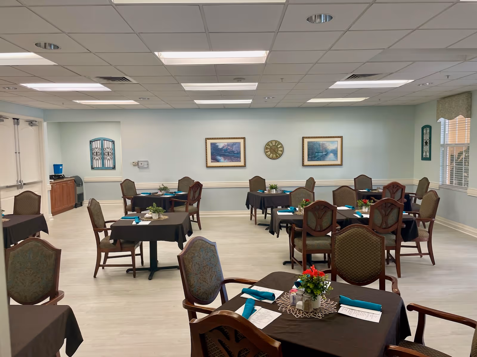 A dining room in an assisted living facility with several tables covered in dark tablecloths, each set with teal napkins, menus, and small floral centerpieces. The room has light-colored walls adorned with framed landscape paintings and a clock. There are multiple chairs around each table, and the floor is light wood. The ceiling has recessed lighting panels.