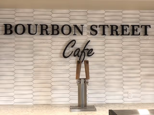 A wall with white hexagonal tiles and black letters spelling 'BOURBON STREET Cafe' above two wooden-handled beverage taps on a countertop.