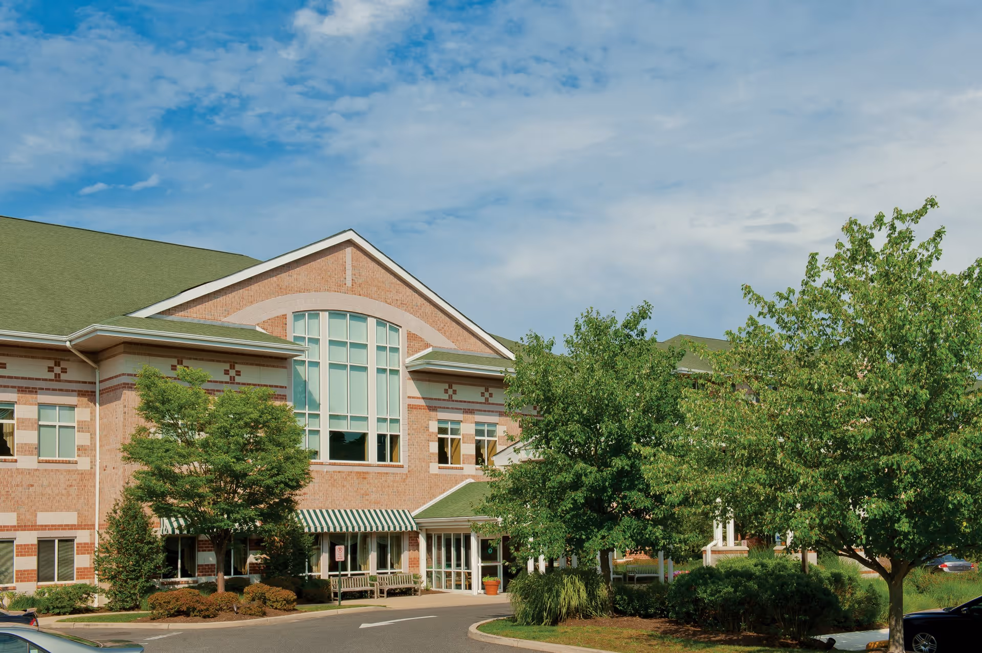 Exterior view of a brick senior living facility building with large windows, a green roof, and a striped green and white awning over the entrance. There are trees and shrubs in front of the building and a clear blue sky with some clouds above.