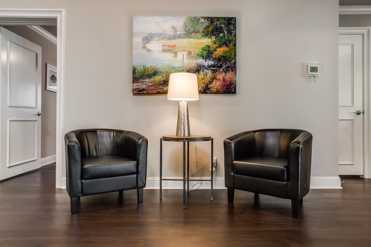 Two black leather armchairs flank a small round table with a lamp beneath a colorful landscape painting in a clean interior seating area.