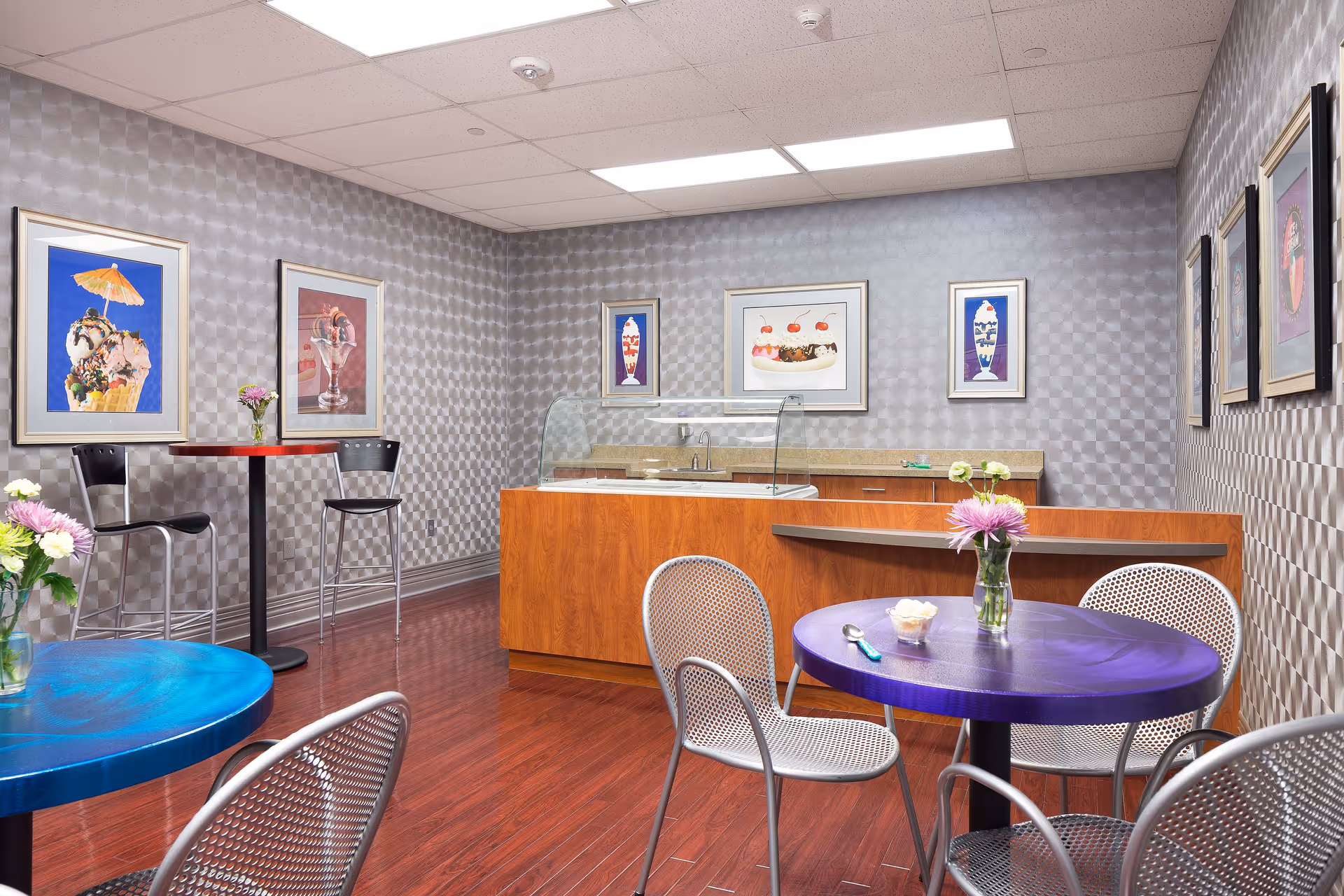 Small dining area with round tables and metal chairs, a wooden service counter, and framed artwork on patterned walls.