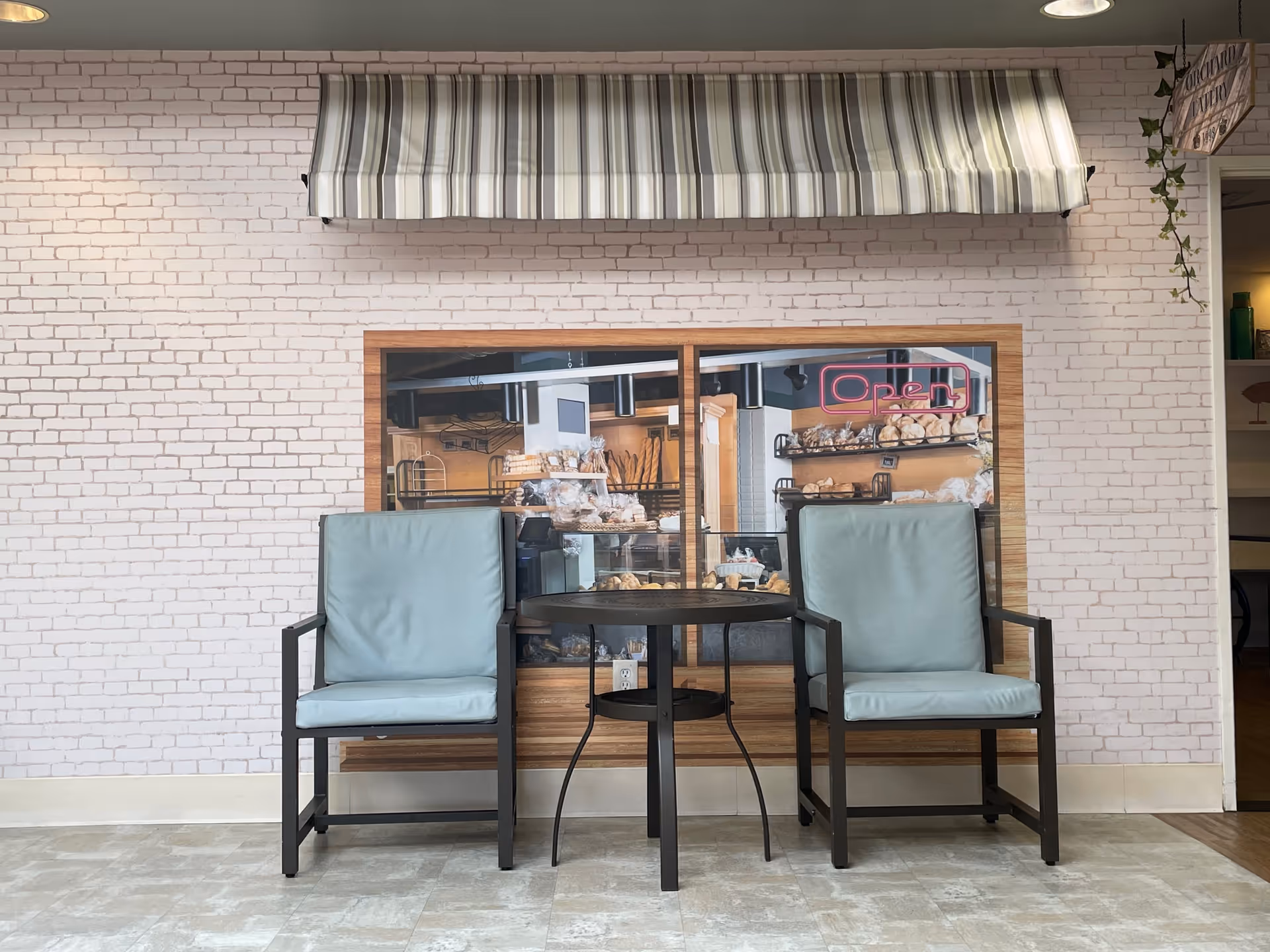 Two light blue cushioned chairs with black metal frames are placed on either side of a small round black table. Behind them is a wall with white brick wallpaper and a large framed picture of a bakery display with bread and pastries, featuring a neon pink 'Open' sign. Above the picture is a striped fabric awning in shades of gray, white, and beige. The floor is tiled in a light beige color.