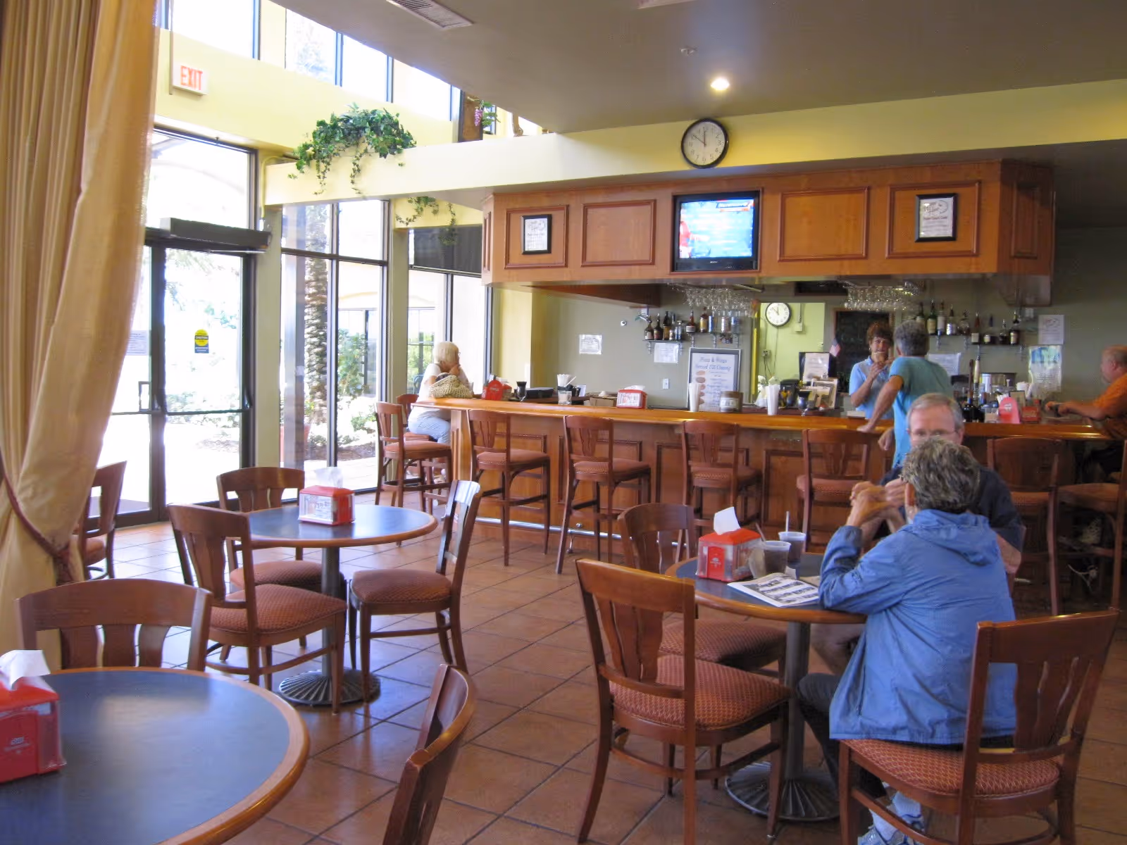 Interior view of a dining area with several round tables and wooden chairs. A few people are seated and conversing. There is a bar counter with bar stools, bottles, and glasses behind it. A clock and a television are mounted above the bar. Large windows and a glass door allow natural light into the room.