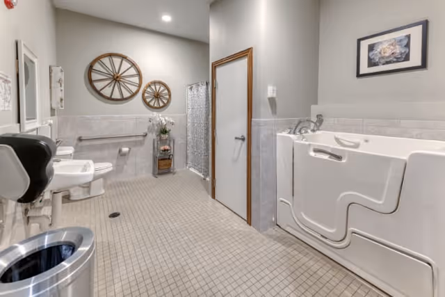 A spacious, accessible bathroom with a walk-in bathtub on the right, a toilet and sink on the left, decorative wall art including two wooden wagon wheels, a small plant stand with flowers, and a tiled floor and walls.