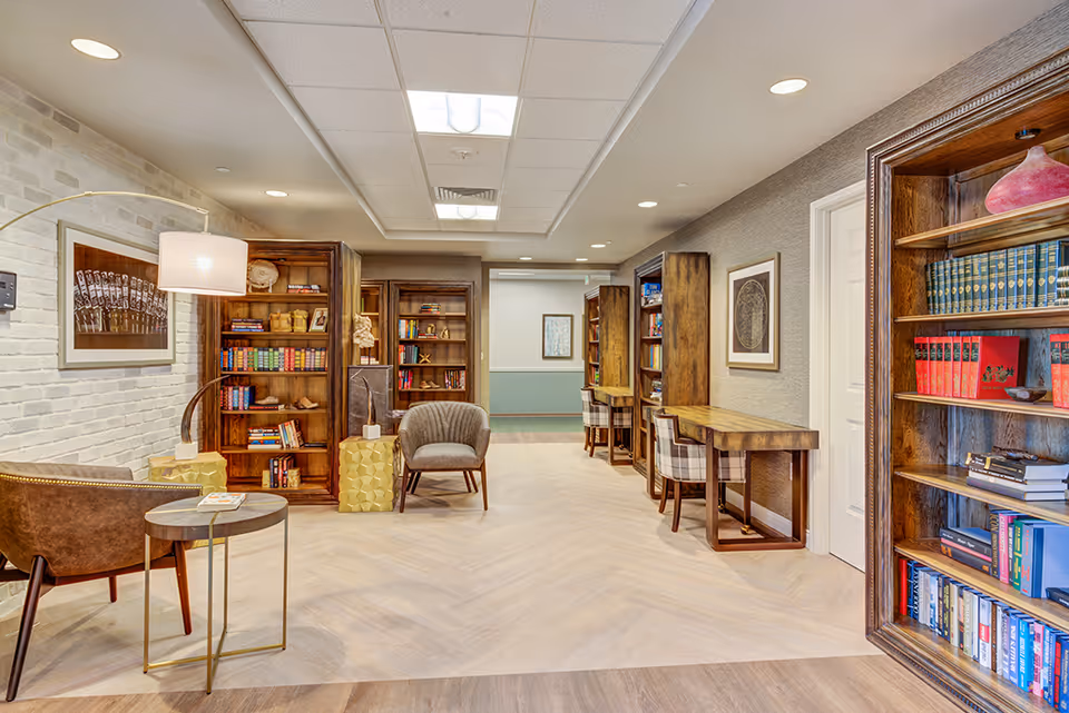 A cozy reading and study area in a senior living facility featuring wooden bookshelves filled with books, comfortable chairs, small tables, and desks with chairs along the wall. The space has warm lighting, a white brick accent wall, and framed artwork.