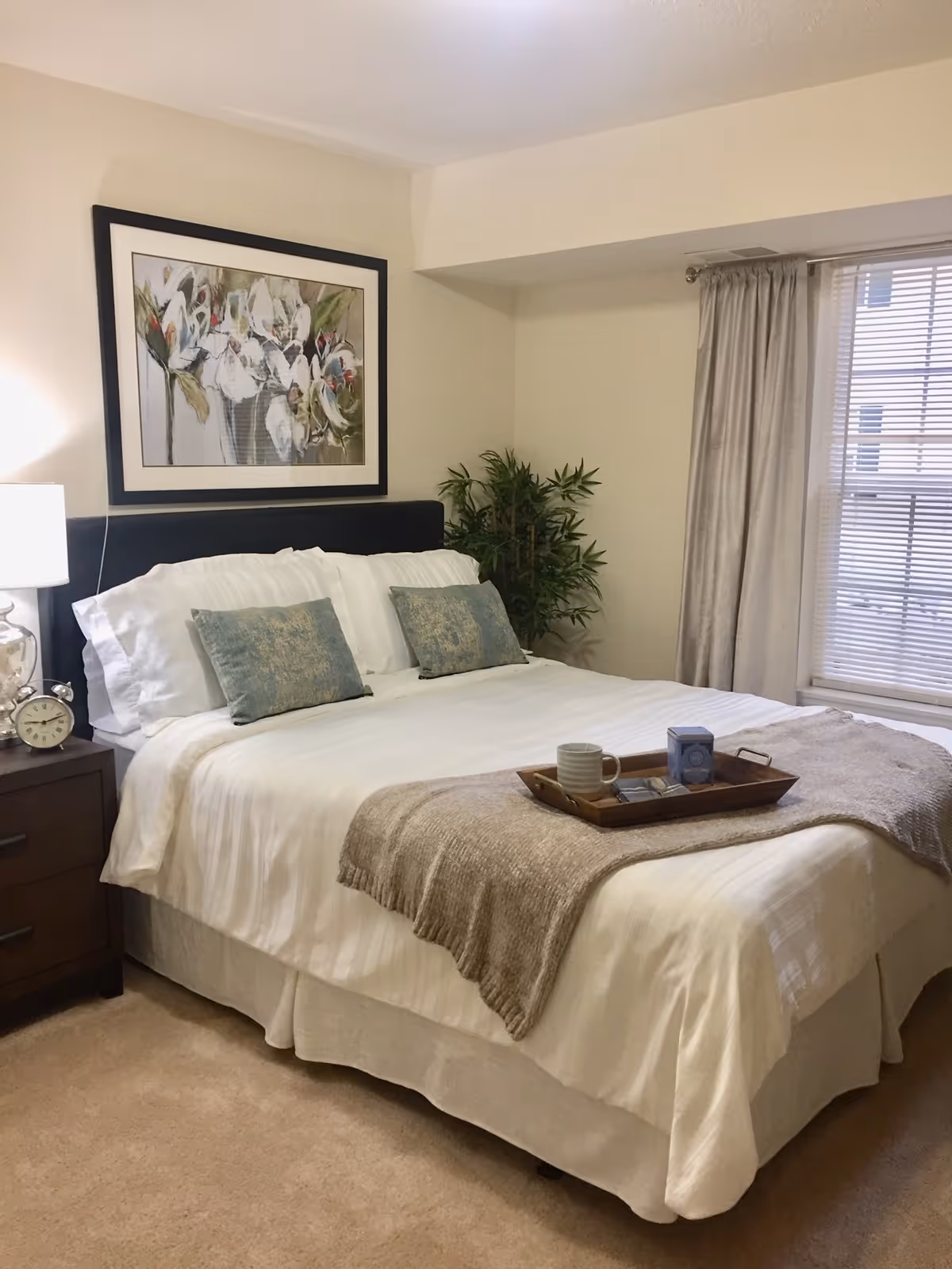 A neatly made bed with white linens, decorative pillows and a tray, beside a nightstand with a lamp, potted plant, framed artwork and a window with blinds.
