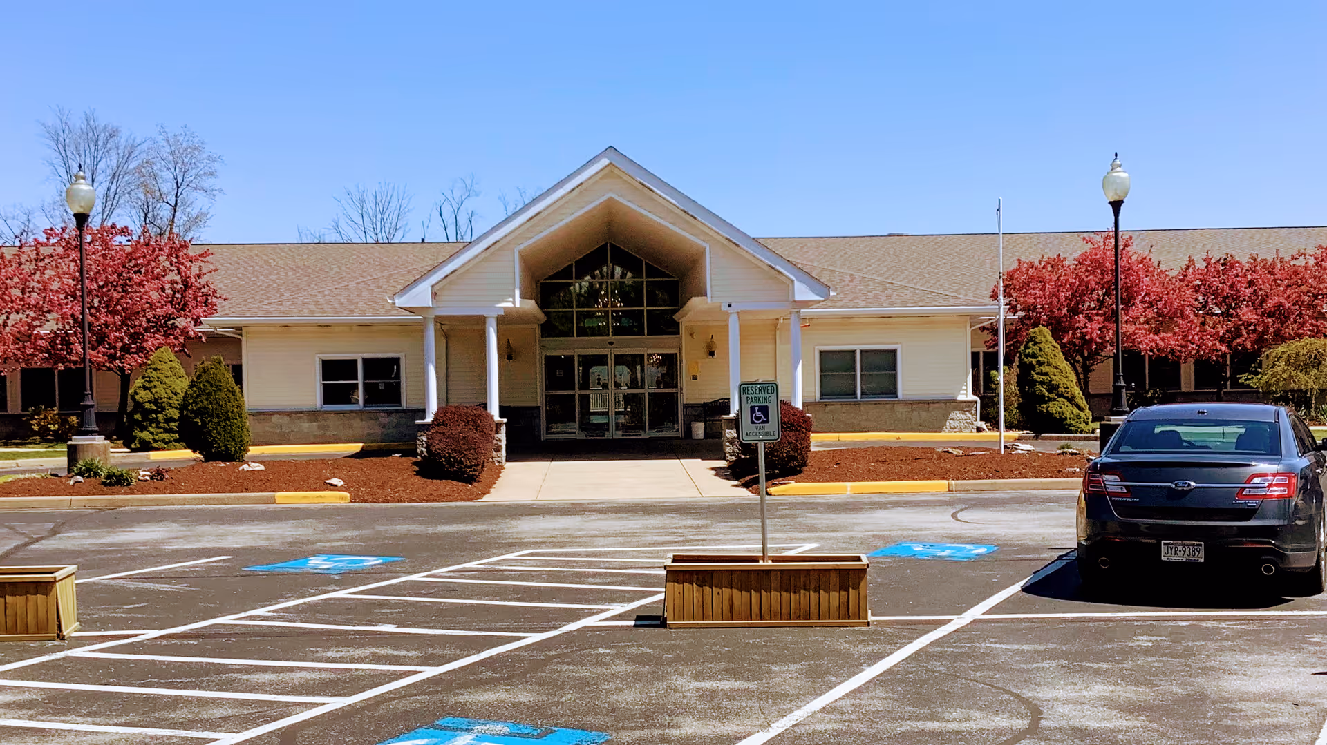 Front entrance of a single-story care facility with a covered portico, landscaped beds, marked handicap parking spaces, and a parked car.
