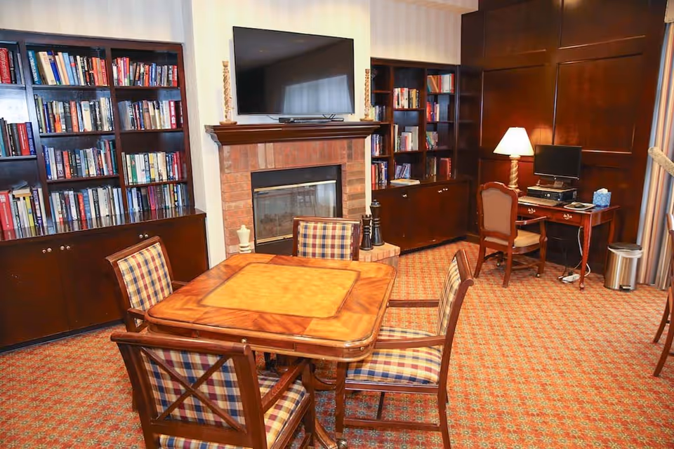 Cozy common room with a wooden card table and plaid chairs, bookshelves, a fireplace with TV above, and a desk with a computer.