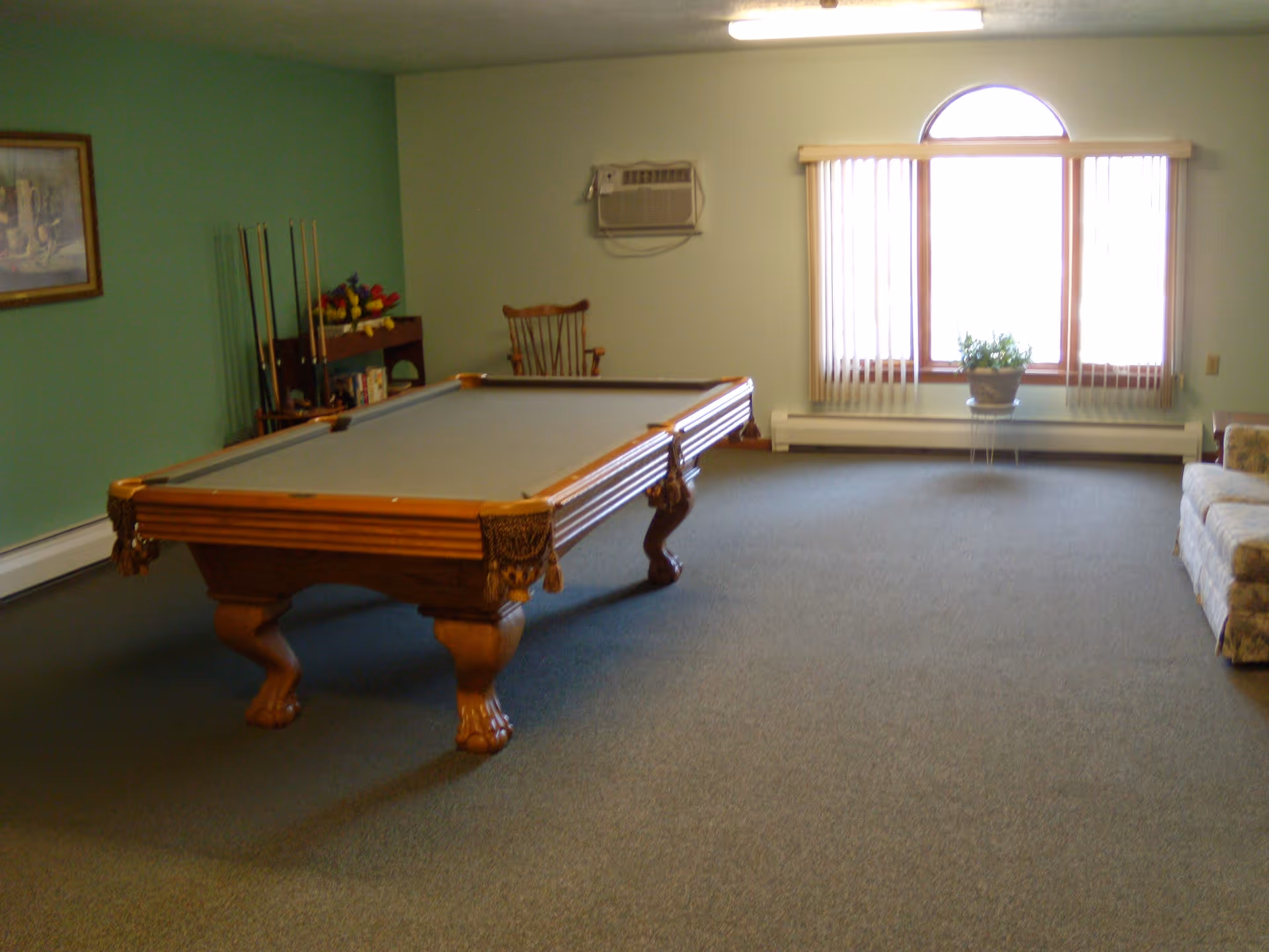 A recreation room with a pool table in the center, a wooden chair and a rack holding pool cues against a green wall. There is a large window with vertical blinds and a potted plant on a small stand in front of it. A floral-patterned couch is partially visible on the right side.