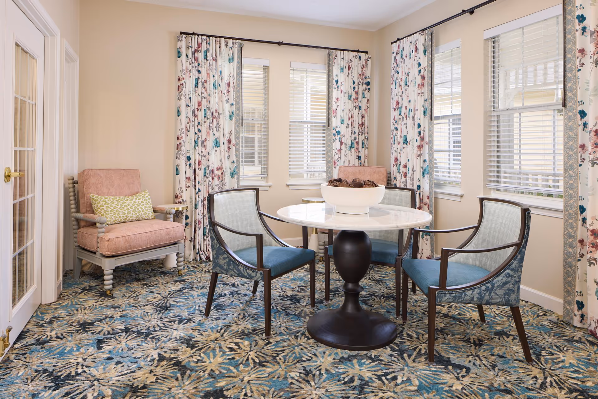 A bright room with a round white marble table surrounded by four blue cushioned chairs with wooden frames. Two pink cushioned armchairs with a green patterned pillow are placed near the windows. The room has large windows with floral curtains and a patterned carpet in shades of blue, beige, and cream.