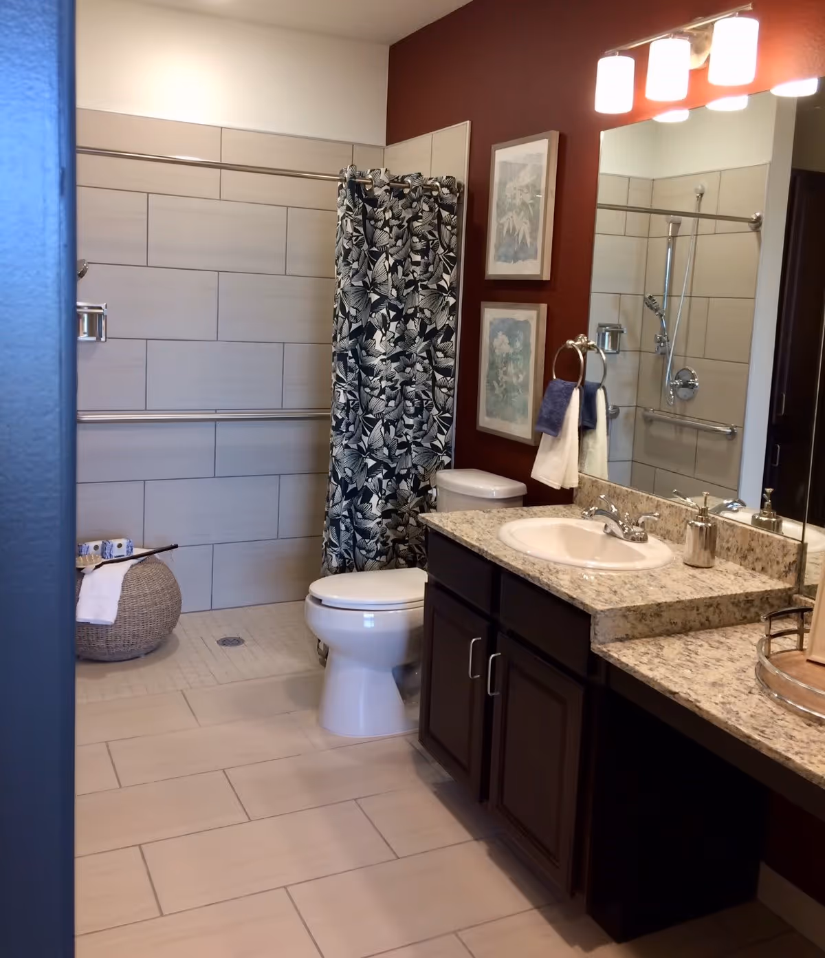 A bathroom with a tiled floor and walls, featuring a toilet, a shower area with a floral patterned shower curtain, a granite countertop with a sink, a large mirror, and two framed pictures on a red accent wall. There are also towels hanging on a ring near the sink and a woven basket with a towel near the shower.