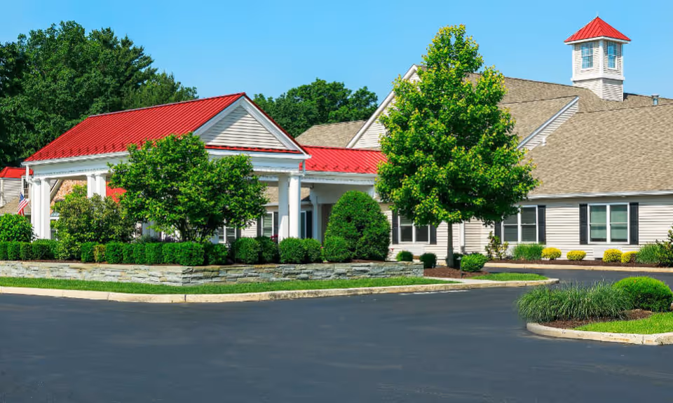 Exterior view of a senior living facility with beige siding and a red metal roof entrance canopy. The building is surrounded by well-maintained landscaping including green bushes, trees, and a stone retaining wall. The sky is clear and blue.
