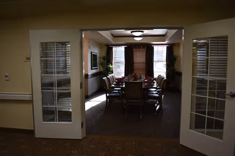 View through open double doors into a dining room with a long table set for a meal, surrounded by chairs. The room has large windows with blinds, plants in the corners, and a ceiling light fixture.