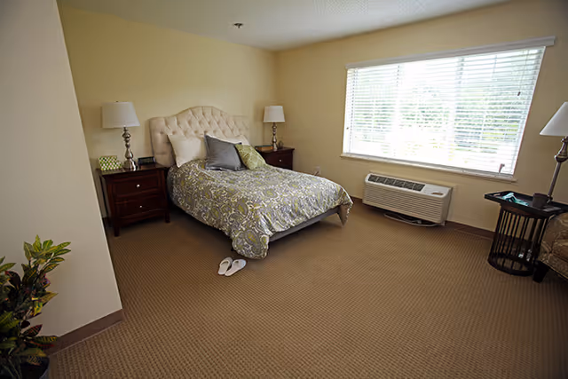A bedroom with a bed featuring a tufted headboard and patterned bedding, two bedside tables with lamps, a window with blinds, an air conditioning unit below the window, a pair of slippers on the carpeted floor, a small table with a chair, and a plant in the corner.