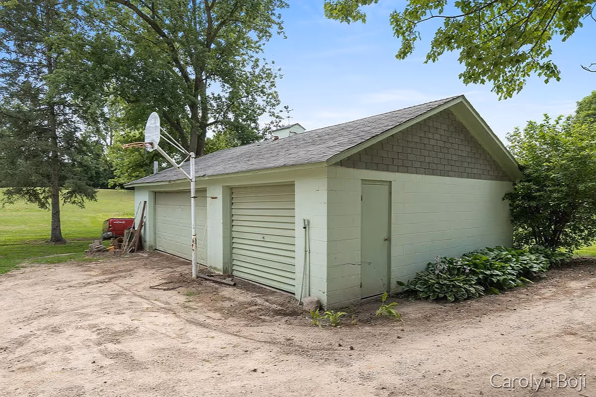 Exterior view of a small detached garage building with two closed garage doors and a side door, surrounded by trees and greenery. There is a basketball hoop attached to a pole in front of the garage, and a dirt driveway leading up to it under a clear blue sky.