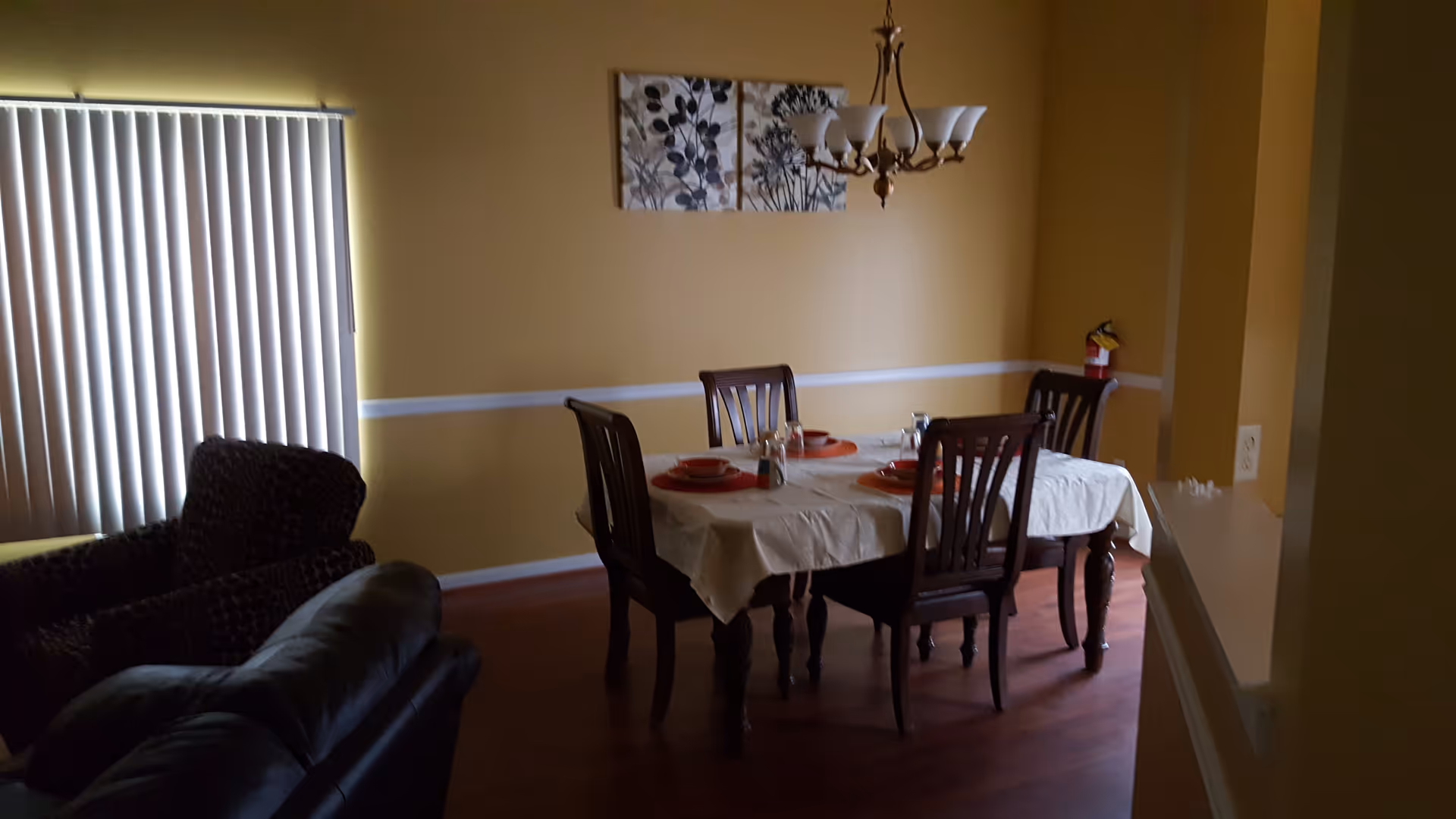 A dining room with a wooden table covered by a white tablecloth, set with four place settings including plates, bowls, and glasses. Four wooden chairs surround the table. A chandelier with five lights hangs above the table. The walls are painted yellow with a white chair rail. There is a window with vertical blinds on the left side, and two armchairs partially visible in the foreground. A fire extinguisher is mounted on the far wall.