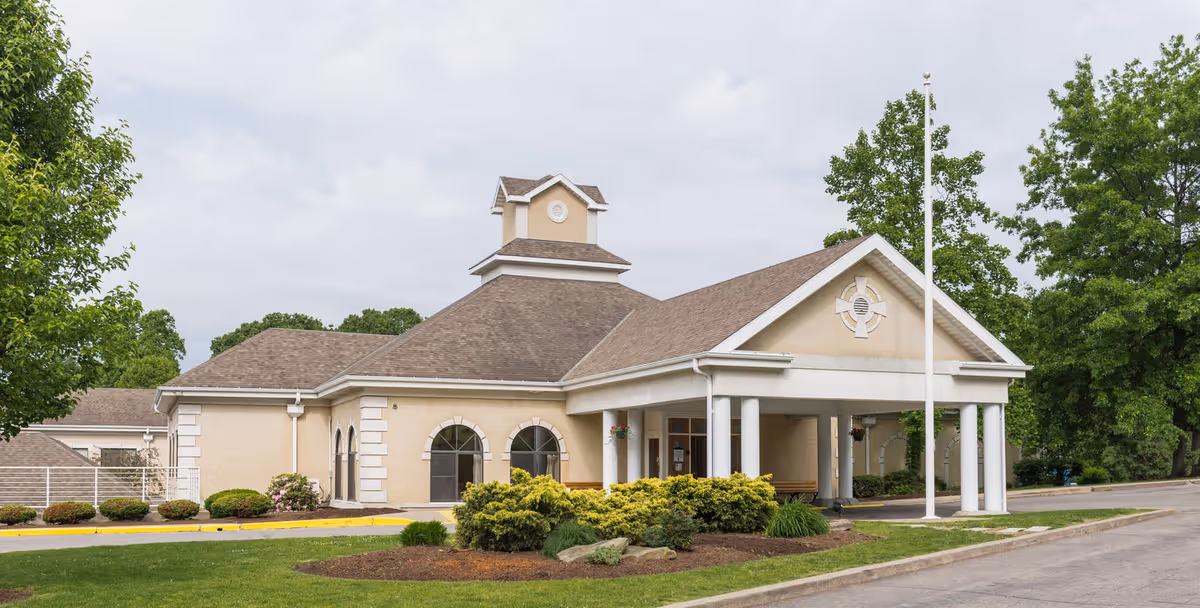 Front entrance of a single-story senior living facility with a covered porte-cochere, flagpole, and landscaped beds.