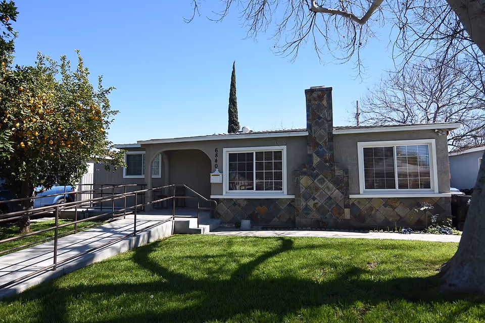 Exterior front view of a single-story senior care facility building with a wheelchair accessible ramp, two large windows with white frames, a stone-tiled chimney, and a green lawn with trees under a clear blue sky.