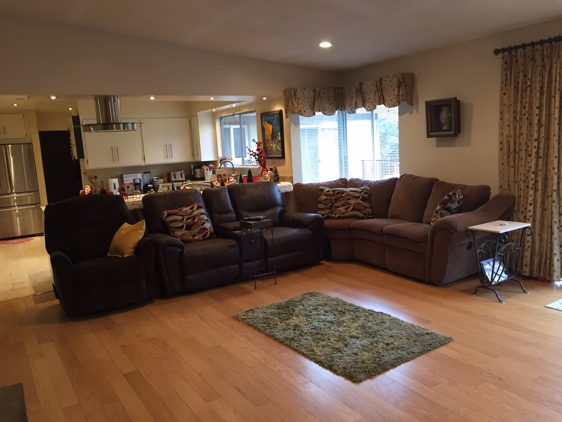 A cozy living room with a brown sectional sofa and a dark recliner chair, decorated with patterned throw pillows. There is a small green rug on the wooden floor in front of the seating area. The room has large windows with beige curtains and valances, allowing natural light to enter. In the background, a kitchen area with white cabinets, stainless steel refrigerator, and various kitchen appliances is visible.
