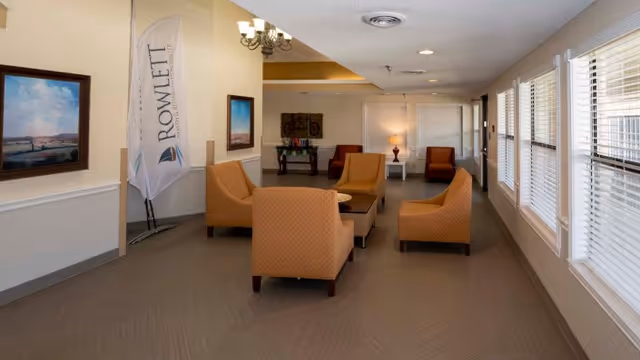 A well-lit common area in Rowlett Health & Rehabilitation Center featuring four orange upholstered chairs arranged around a rectangular coffee table. The room has large windows with blinds on the right side, two framed landscape paintings on the left wall, a standing flag with the facility's name, and additional seating with two orange chairs and a small table with a lamp in the background.
