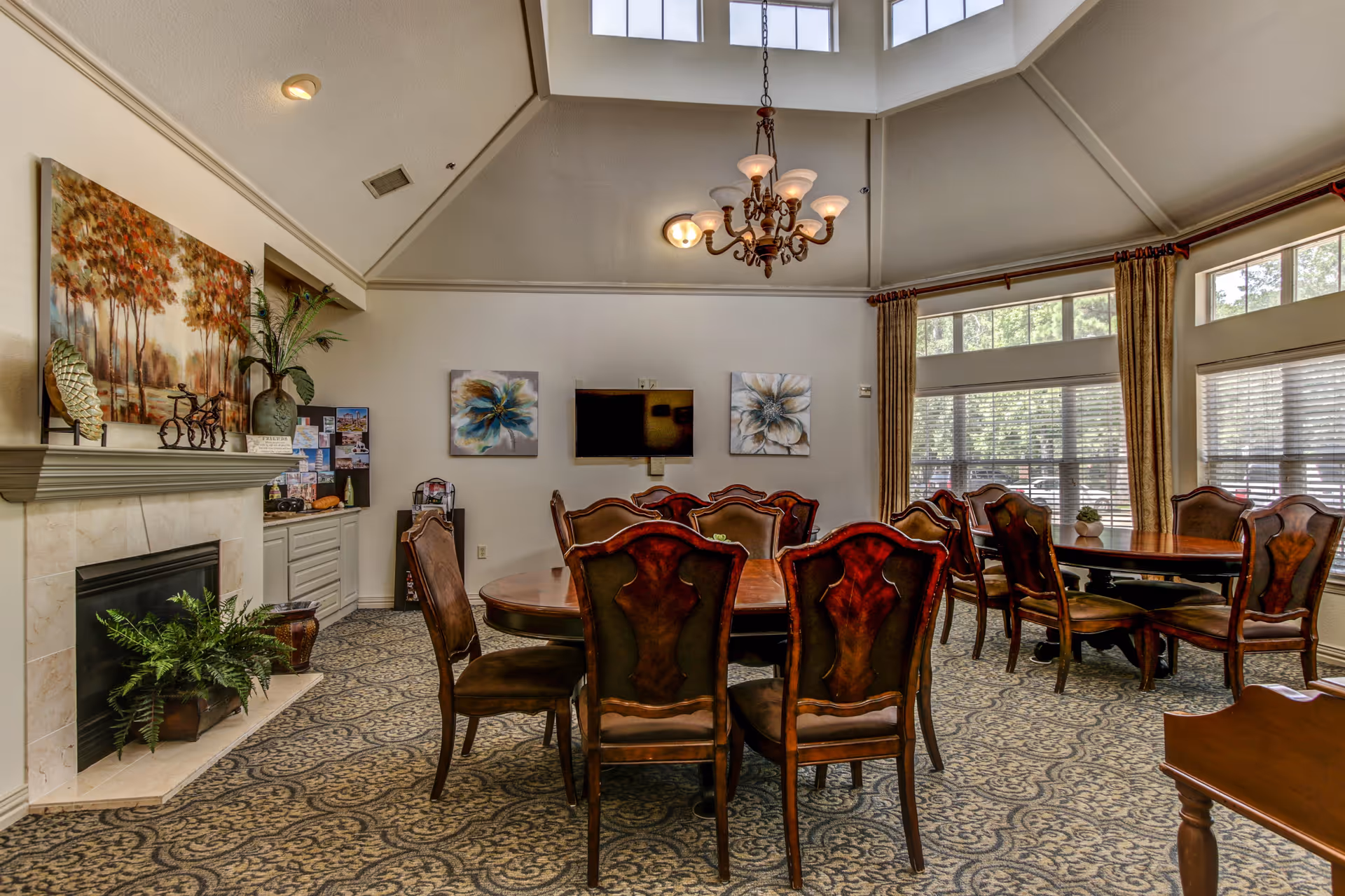 Sunlit senior living common room with several wooden dining tables and chairs, a fireplace, wall art, and large windows.