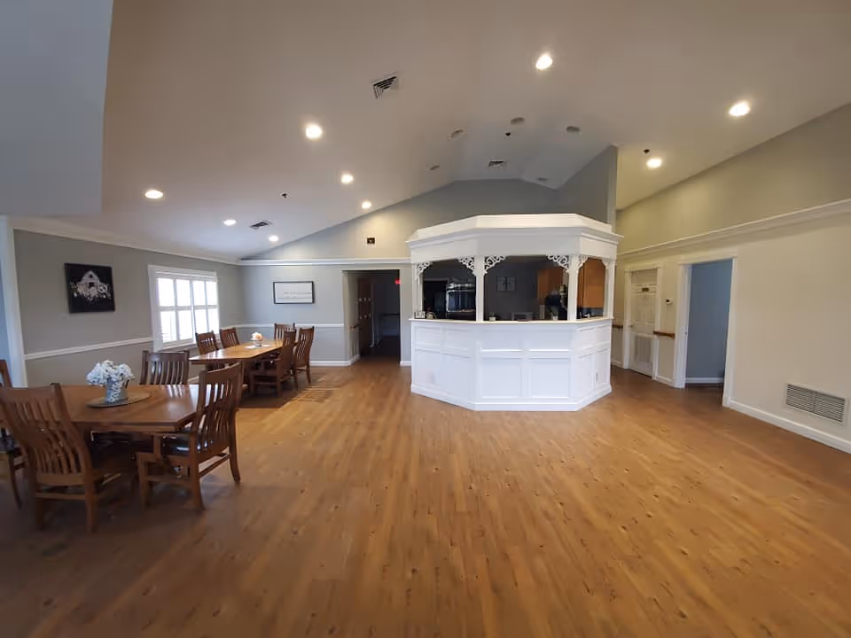 A spacious room with wooden flooring and light gray walls featuring a white octagonal reception desk or counter in the center-right. To the left, there are two wooden dining tables with chairs, each table decorated with a small flower arrangement. The room has recessed ceiling lights and a window with white shutters on the far left wall. There are two doorways visible, one in the center and one on the right side of the room.