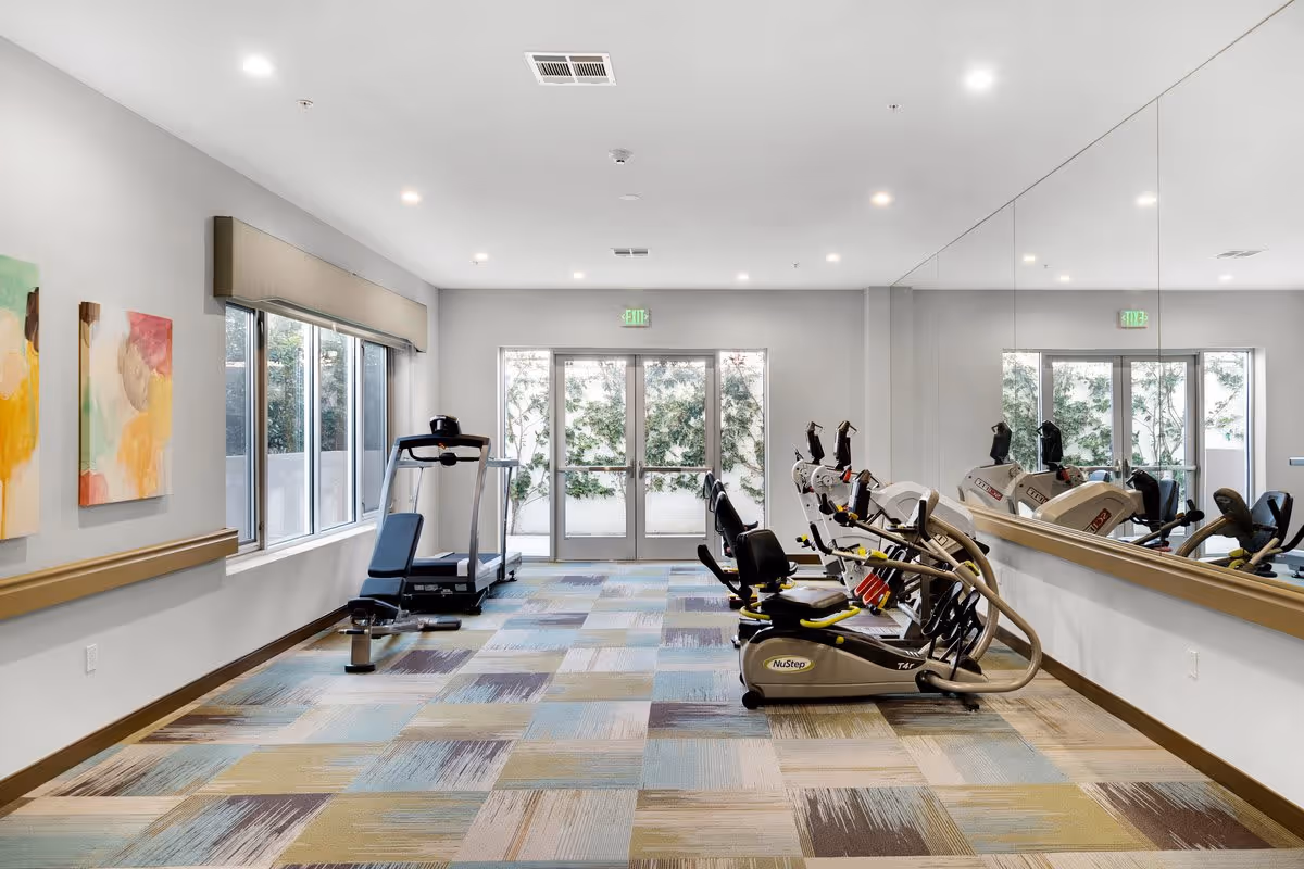 A bright and clean exercise room in a senior living facility with large windows on the left, colorful abstract paintings on the wall, and a mirrored wall on the right. The room contains exercise equipment including a treadmill and several NuStep recumbent cross trainers arranged along the right side.
