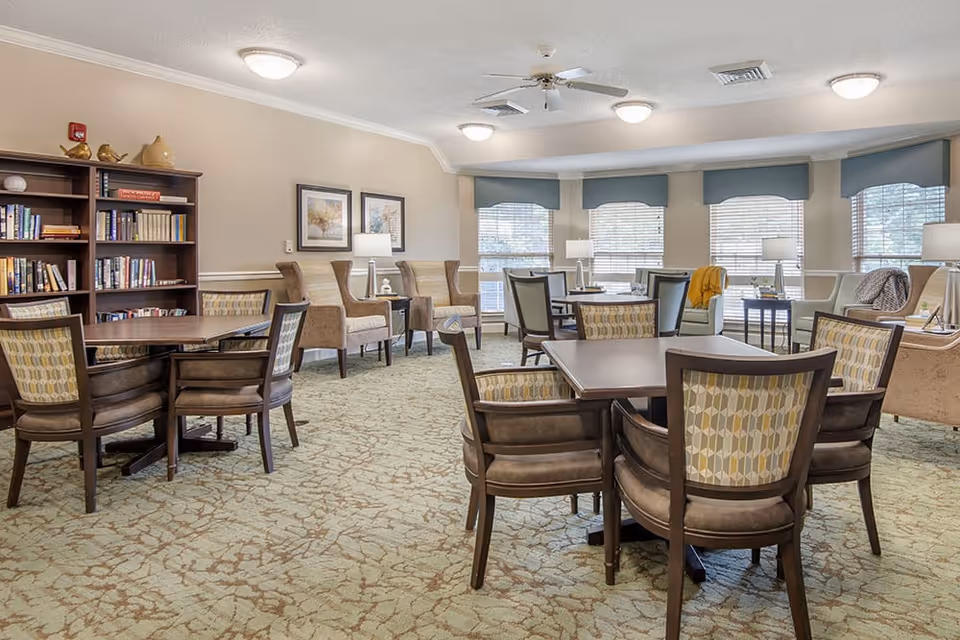 A well-lit common area with multiple tables and chairs arranged for seating. There are several armchairs along the walls, a bookshelf filled with books, and large windows with blinds letting in natural light. The room has a carpeted floor and neutral-colored walls with framed artwork and table lamps.
