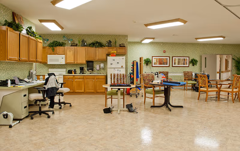A spacious room with a kitchenette featuring wooden cabinets, a microwave, coffee maker, and refrigerator. The room has several tables and chairs arranged for activities, with some equipment and games on the tables. The walls are decorated with framed pictures and plants, and there are office chairs and a desk on the left side.