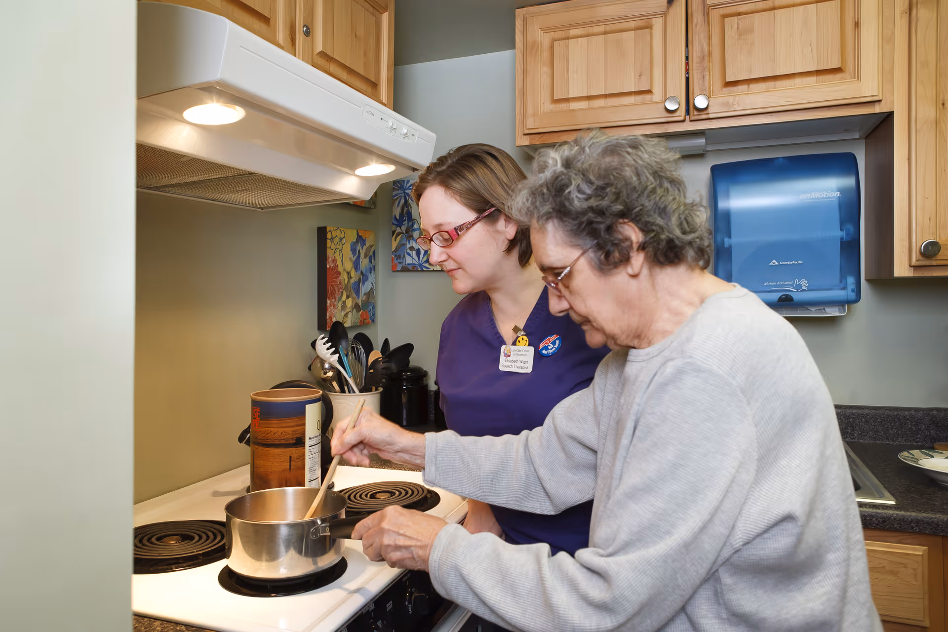 An elderly woman stirring a pot on a stove in a kitchen while a caregiver in a purple uniform watches and assists her. The kitchen has wooden cabinets, a paper towel dispenser, and various kitchen utensils on the counter.