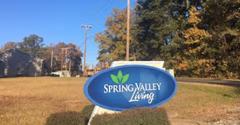 Blue oval Spring Valley Living sign on a grassy lawn with autumn trees.
