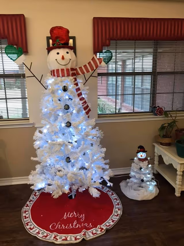 A festive indoor scene featuring two white Christmas trees decorated as snowmen with red hats, scarves, and green mittens. The larger tree is adorned with white lights and black buttons, standing on a red tree skirt that reads 'Merry Christmas.' The smaller tree also has white lights and similar decorations. The background shows windows with blinds and a small table with potted plants.