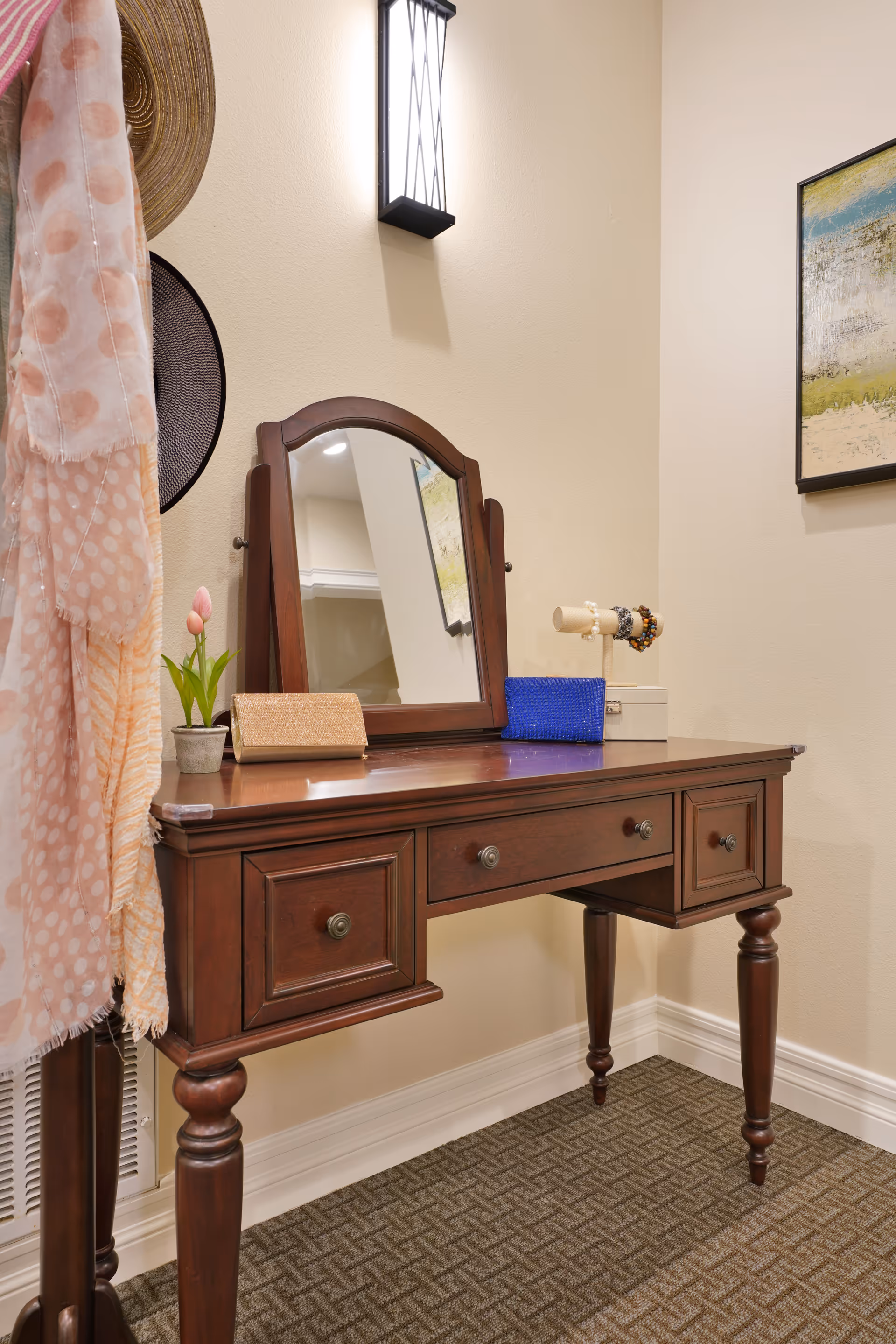 A wooden vanity table with an attached mirror in a corner of a room. The table has three drawers and is decorated with a small potted plant, a gold clutch, a blue clutch, and a jewelry holder with bracelets. A wall-mounted light fixture is above the mirror, and a framed abstract painting hangs on the adjacent wall. A pink and white polka dot scarf and a hat hang on a stand next to the vanity.