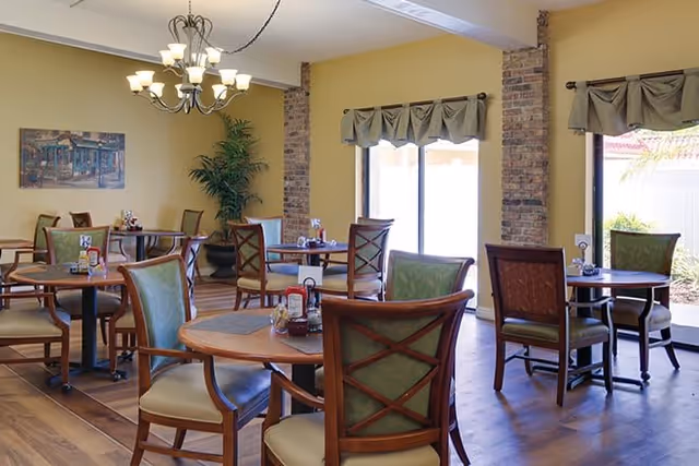 A bright dining room with several round wooden tables and cushioned chairs arranged neatly. The room has yellow walls with exposed brick columns, large windows with green valances letting in natural light, a chandelier hanging from the ceiling, and a potted plant in the corner. There is a painting on one wall and condiments on the tables.