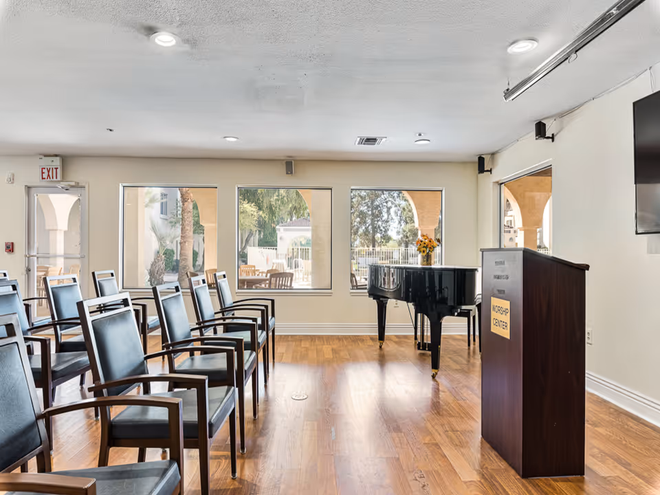 A bright room with wooden flooring featuring rows of black chairs with wooden armrests arranged facing a wooden podium labeled 'Worship Center'. A black grand piano with a vase of flowers on top is positioned near three large windows showing an outdoor patio area with trees and seating. The room has a white ceiling with recessed lighting and an exit door on the left side.