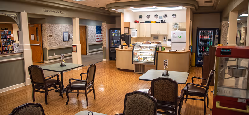Interior view of a small café area named Crossroads Cafe with several tables and chairs arranged on a wooden floor. The café counter displays a variety of food items in a glass case, with a refrigerator and vending machines in the background. The setting appears clean and welcoming with soft lighting.