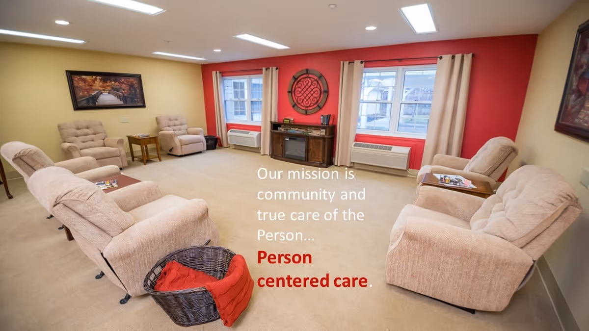 A cozy living room with beige recliner chairs arranged in a semi-circle around a small wooden cabinet with a decorative wall piece above it. The walls are painted yellow and red, with two windows covered by beige curtains letting in natural light. A wicker basket with a red blanket is on the floor, and framed artwork hangs on the walls. Text on the image reads: 'Our mission is community and true care of the Person... Person centered care.'