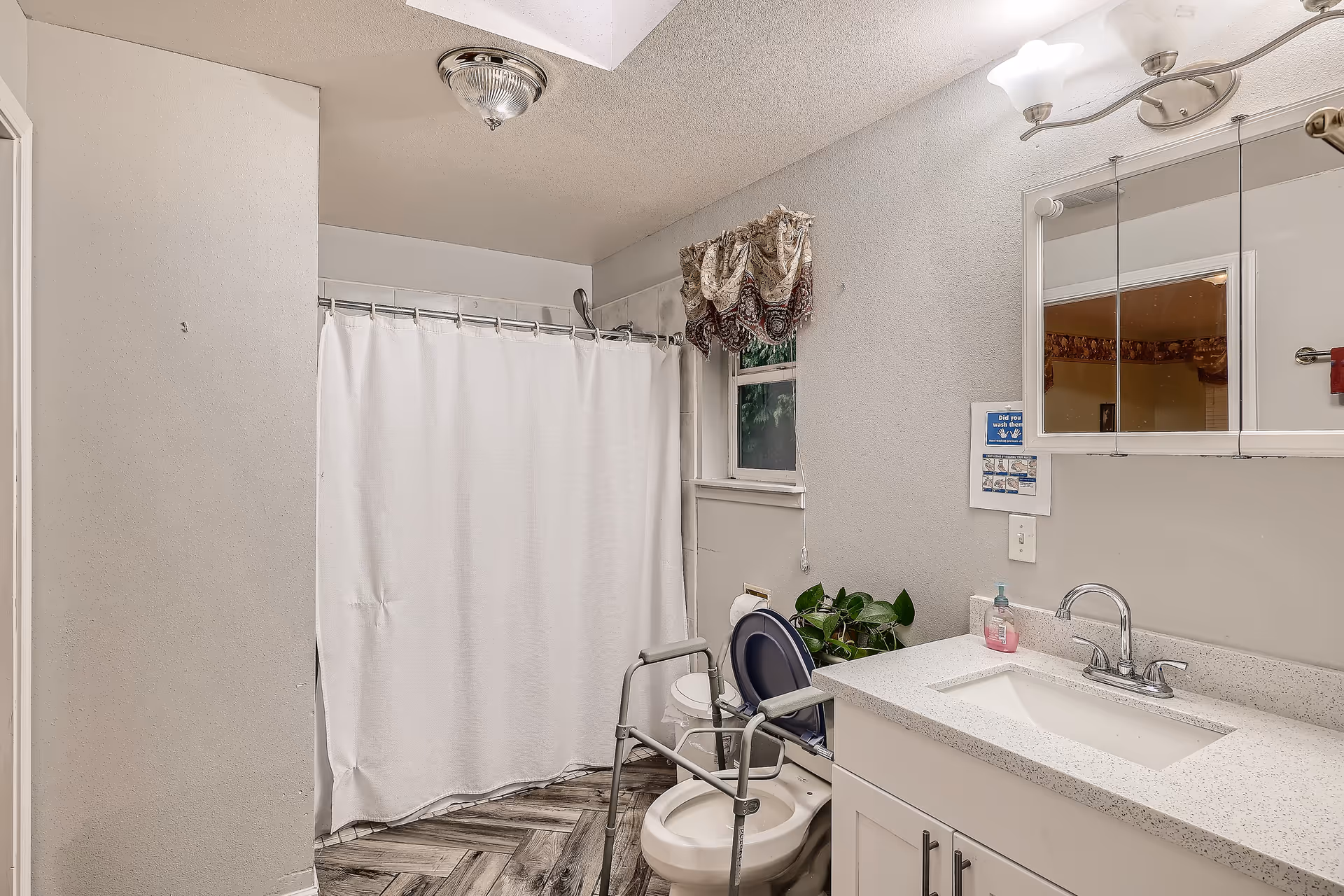 Bathroom with a white shower curtain, a toilet equipped with a safety frame, a white vanity with a sink and faucet, a mirror cabinet above the sink, a small window with a decorative curtain, and a potted plant near the toilet.